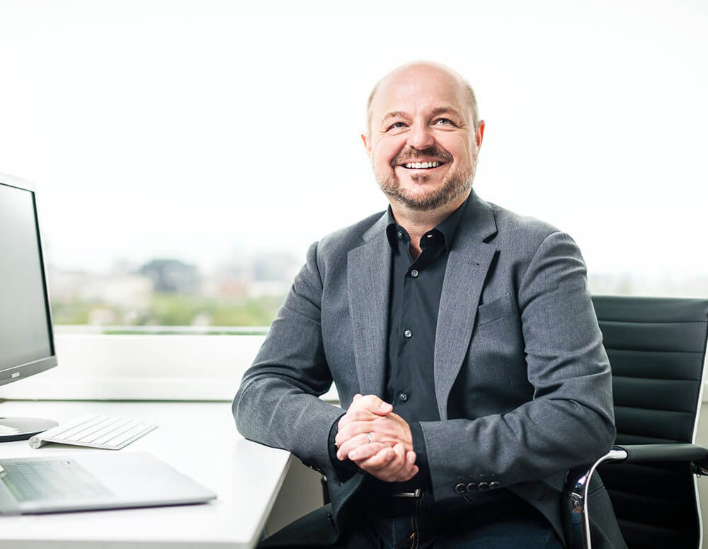 A beaming man in a dark blazer seated in an office setting.