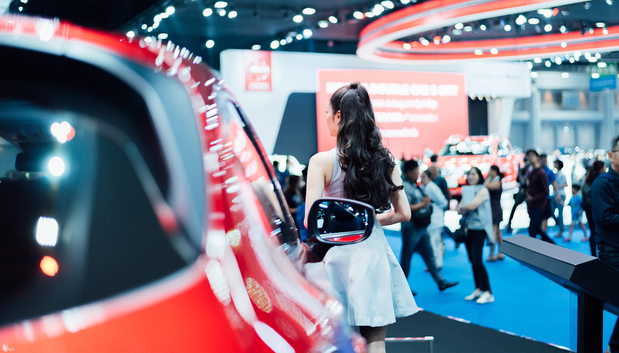 A woman at a car show with a red car in the foreground and event attendees in the background.