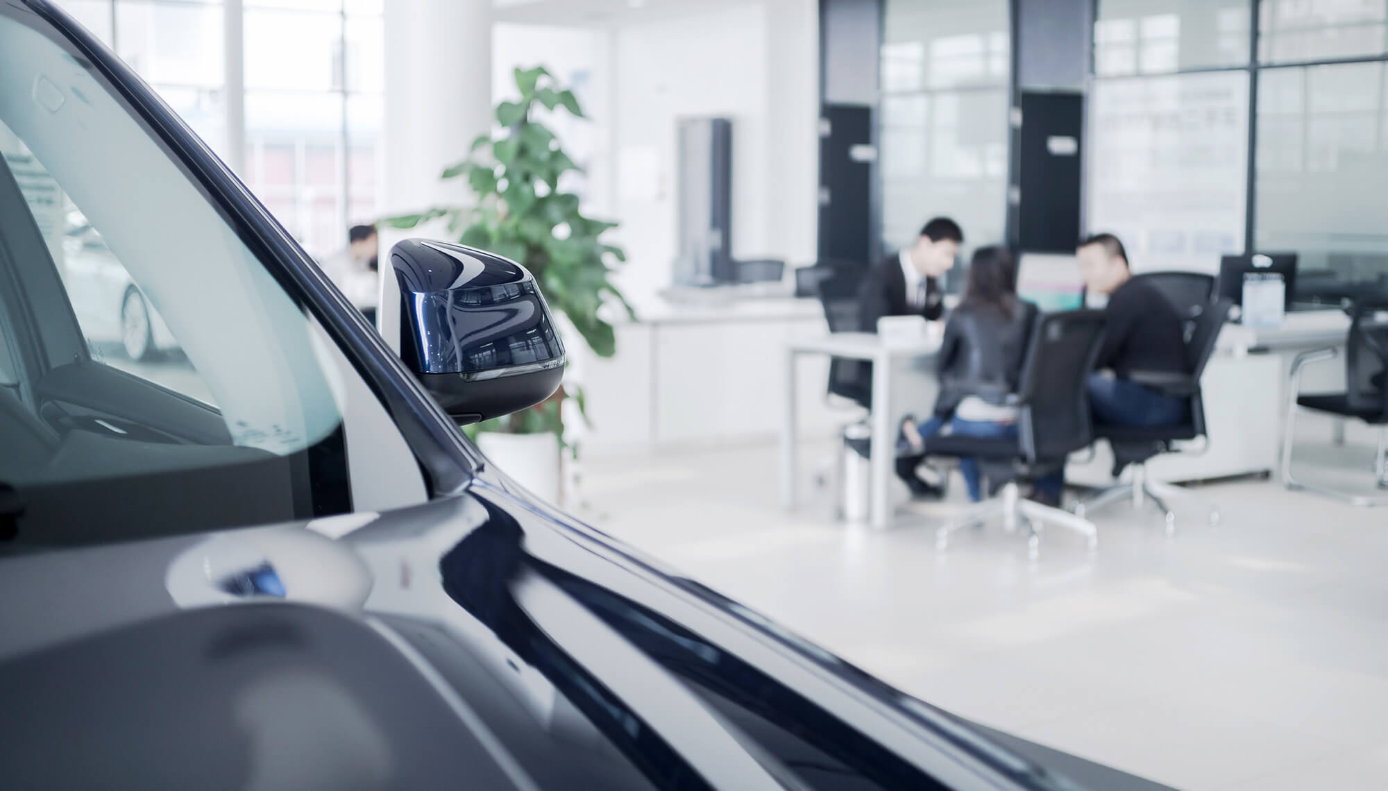 A couple at a car dealership discussing options with a sales representative with a vehicle visible in the foreground.