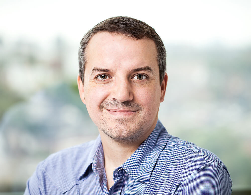 A man in a checkered shirt smiling with a city view in the background.