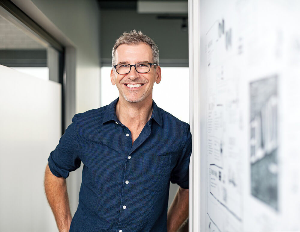 A cheerful man in a dark blue shirt leaning on a doorframe in an office.