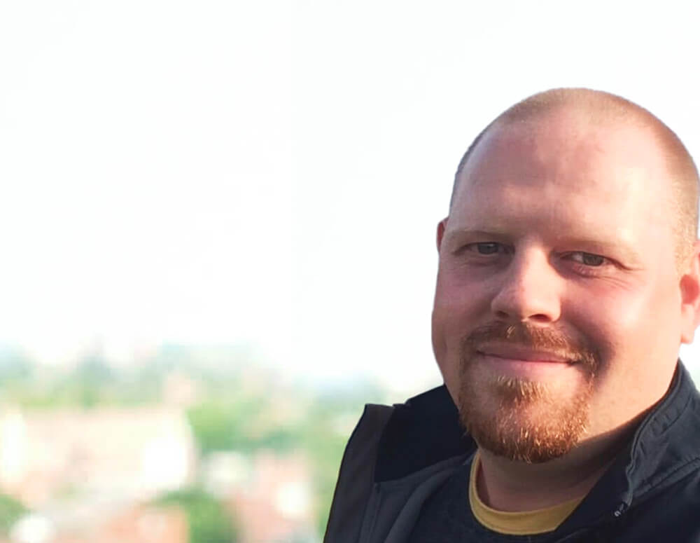 A smiling man with a goatee outdoors with a blurred cityscape.